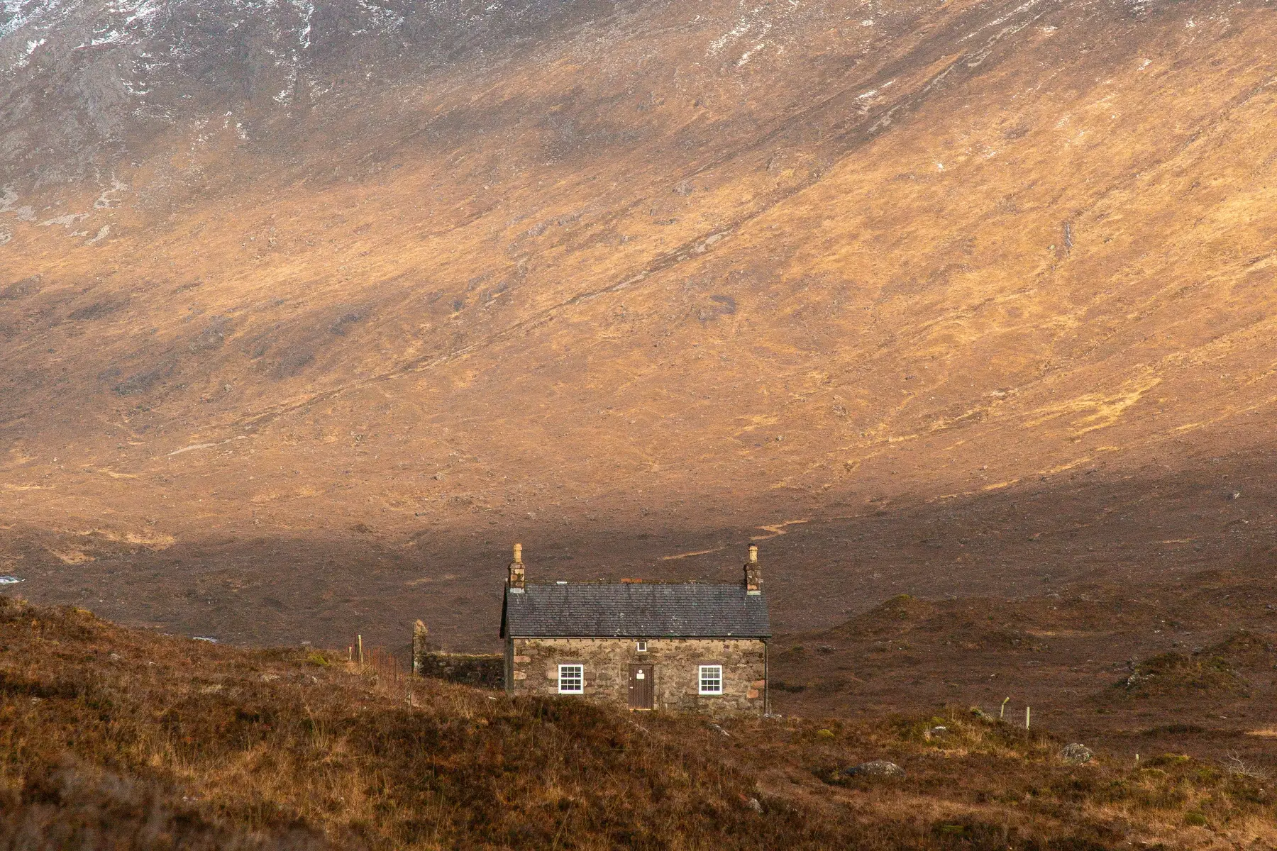 A stone cottage standing alone on open moorland beneath a Highland hillside